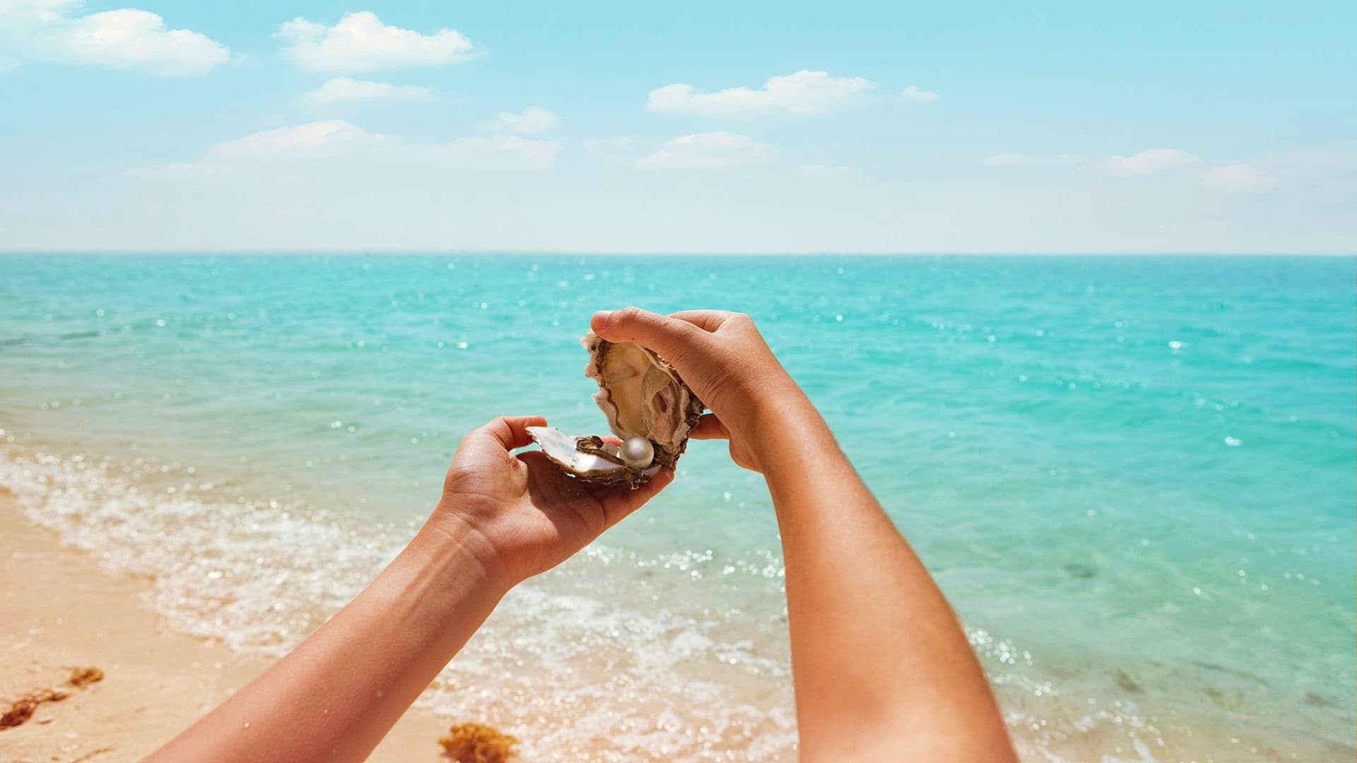 Person holding open oyster shell with pearl against turquoise ocean and sandy beach background